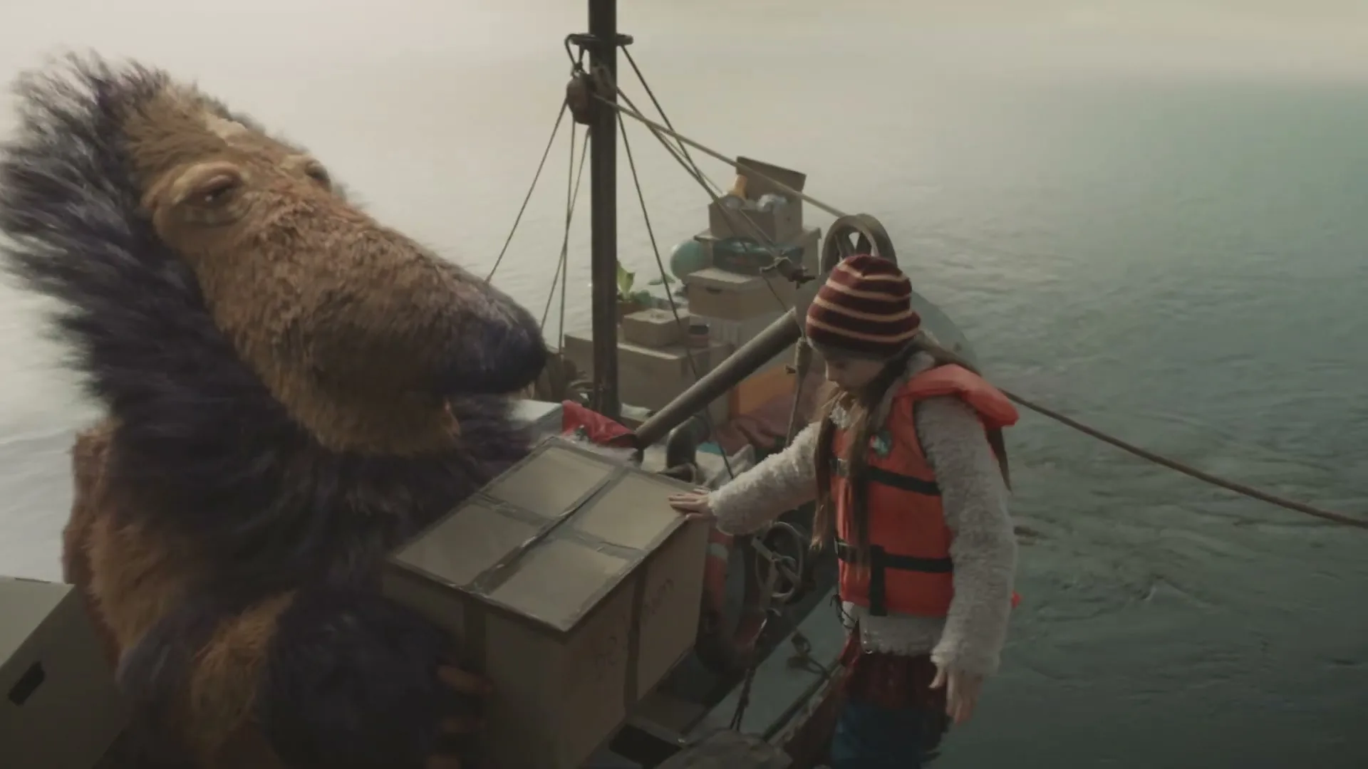 A young girl in a life vest and a large, furry creature with a long snout are on a boat, transferring a cardboard box on a misty body of water.