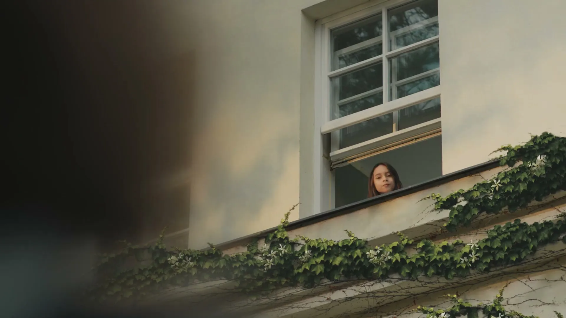A young girl with long brown hair looks out of an open window of a white building covered in ivy, smiling.