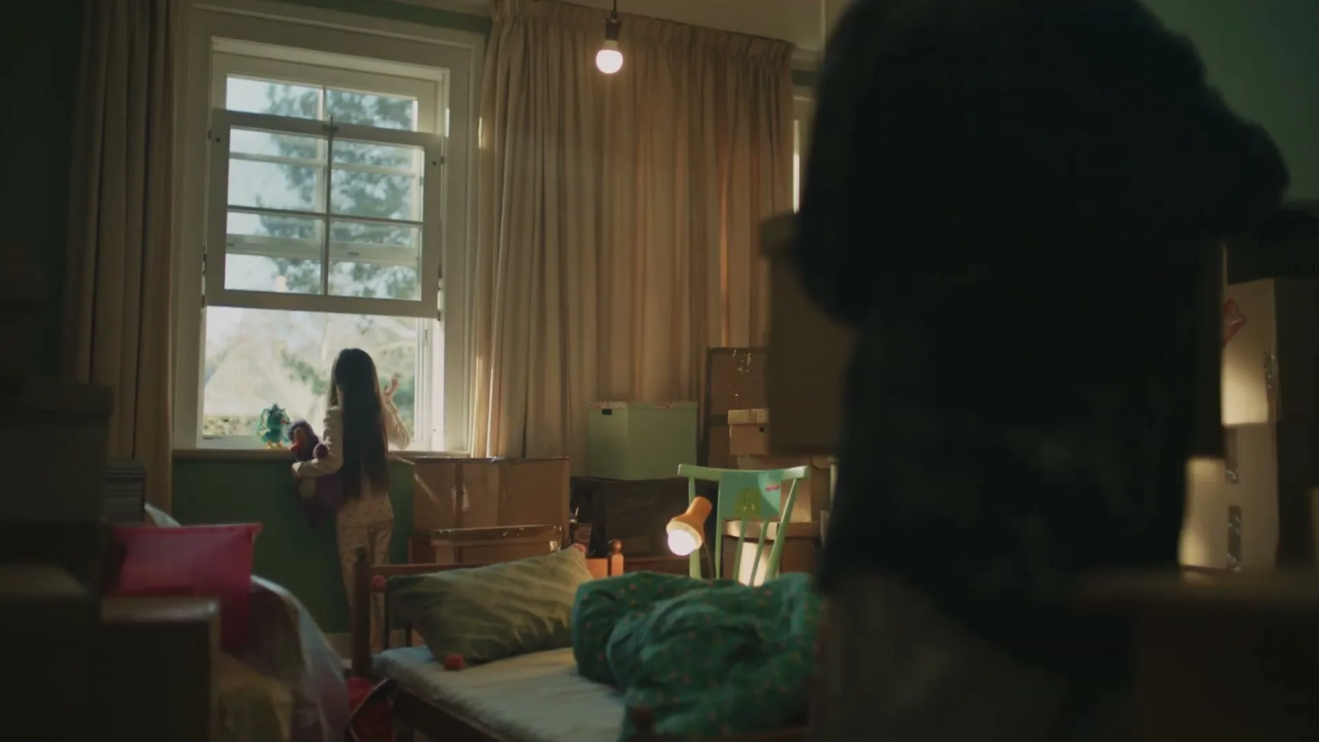 A young girl in pajamas stands by a window, holding a doll, as a man walks past in a room filled with moving boxes.