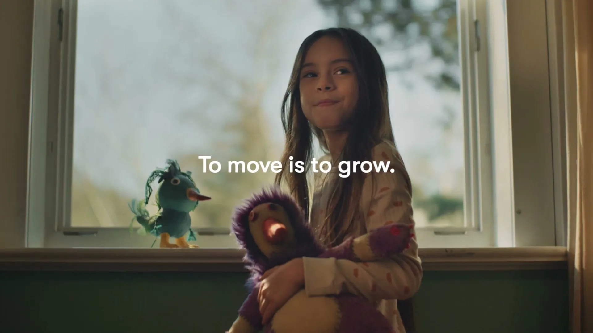 A young girl with long dark hair, wearing pajamas, holds a purple stuffed animal while looking out a window with a blue bird toy on the sill.