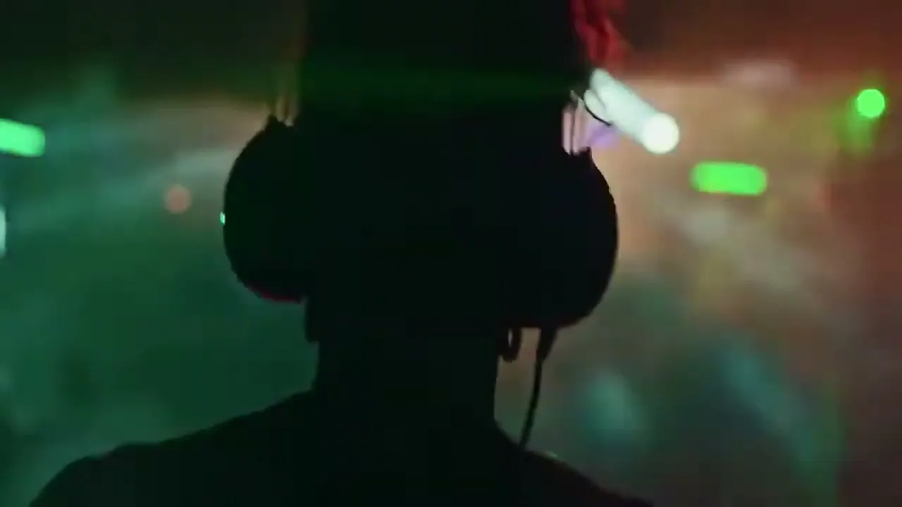 A person with headphones on, silhouetted against bright green and red club lights, is seen from behind in a dark club.
