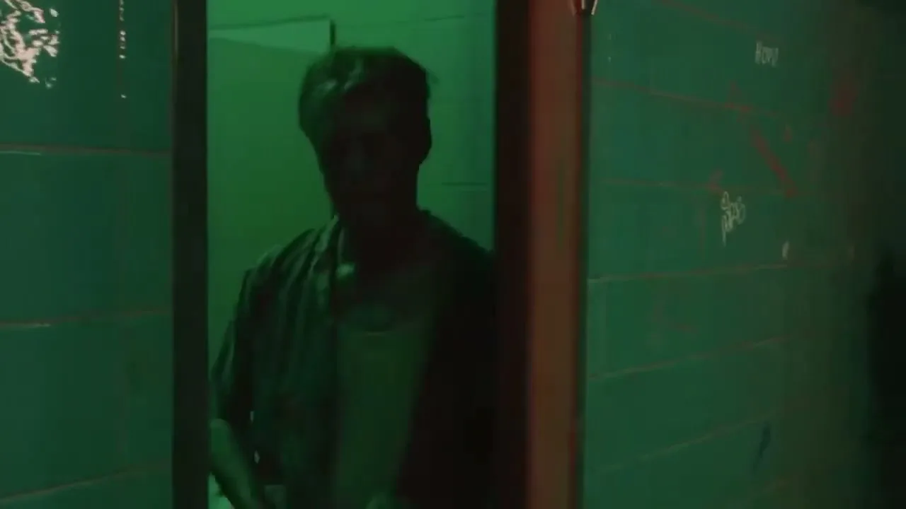 A man with a colorful shirt and white undershirt exits a red door in a green-tiled, graffiti-covered public restroom.