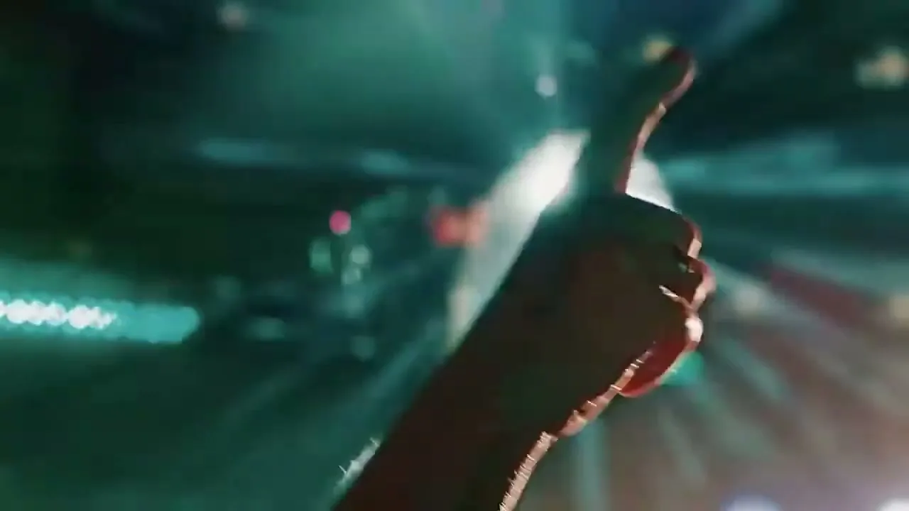 A person's hand with red nail polish points upwards in a dark, blurry club with green and white lights.