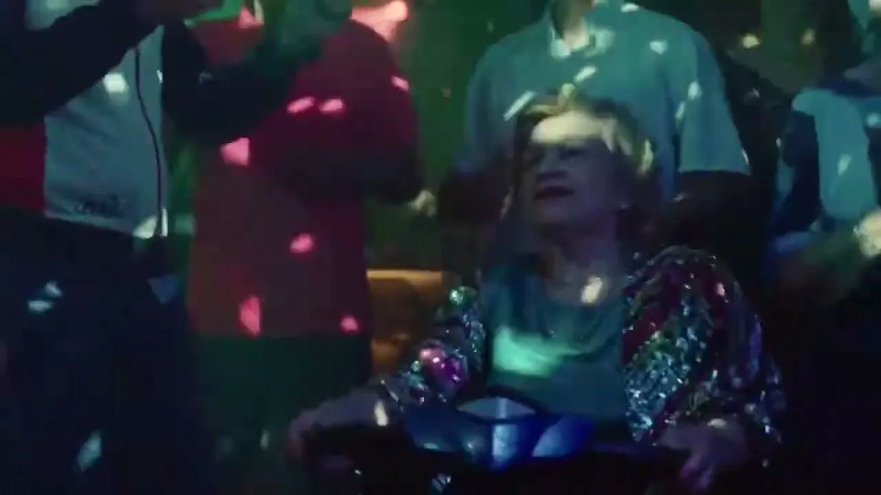 An elderly woman in a sequined jacket sits in a mobility scooter, smiling and dancing amidst a crowd of people in a dimly lit club with disco ball lights.