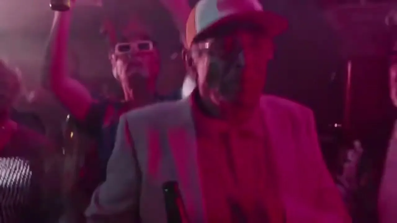 An older man in a cap and suit jacket dances in a crowded, neon-lit nightclub, surrounded by other revelers.