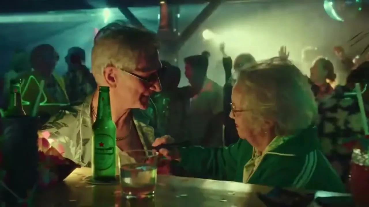 An older man and woman, both with white hair, are at a lively bar, with the man holding a Heineken bottle and the woman showing him something on her phone.