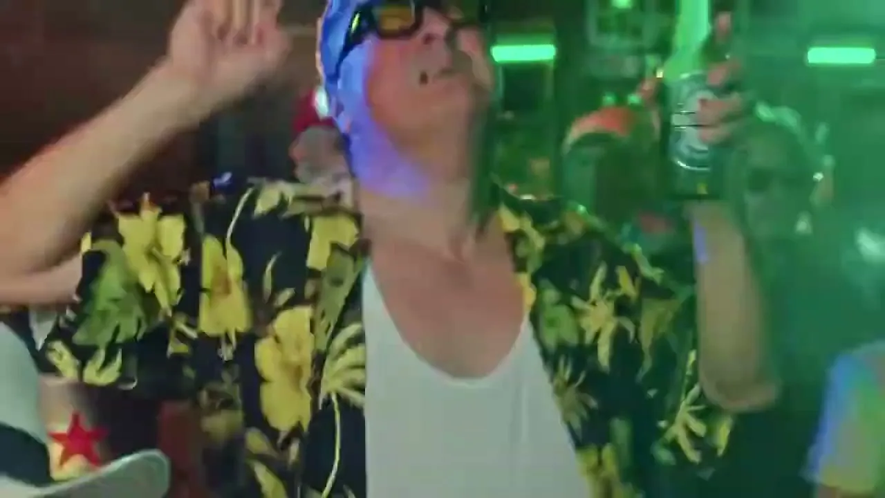 A man in a Hawaiian shirt and sunglasses dances energetically, holding a green bottle, surrounded by other people in a dimly lit club.