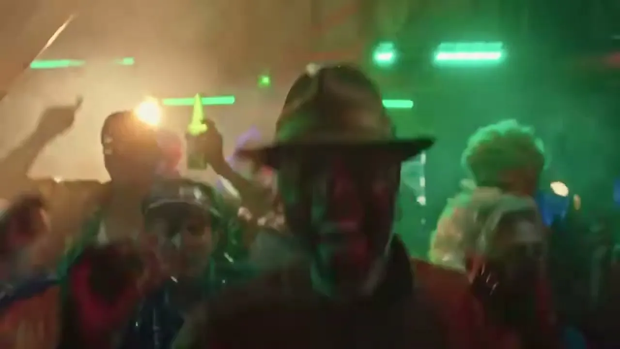 A man in a fedora and gold jacket dances energetically, surrounded by other people in a vibrant, smoke-filled nightclub.