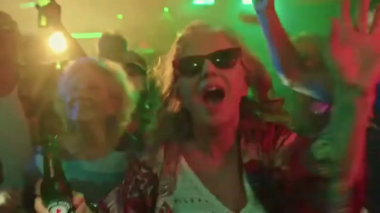 A woman in sunglasses and a patterned shirt screams excitedly while holding a Heineken bottle and dancing in a crowded, neon-lit club.