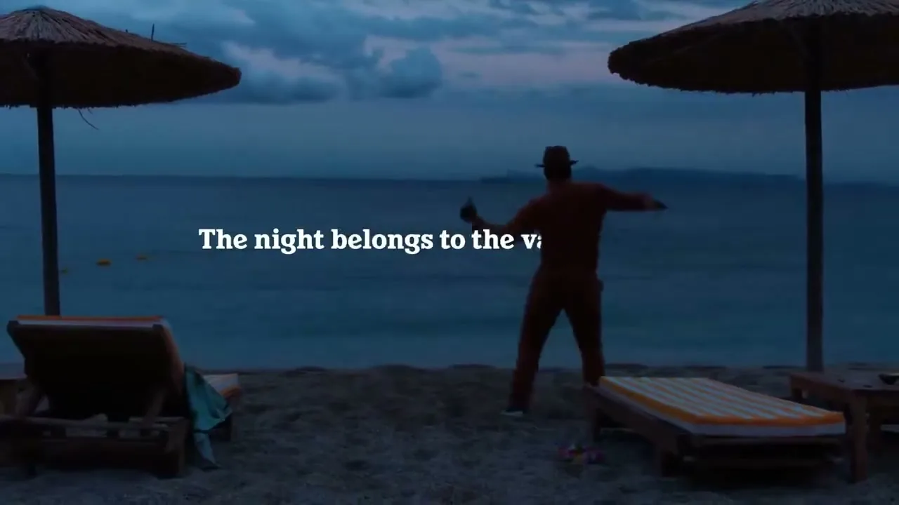 A man in a hat and brown outfit dances on a beach at dusk, with the ocean and distant land in the background, and beach chairs in the foreground.