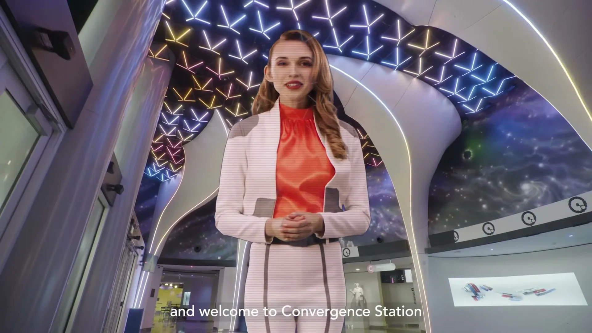 A holographic woman in a futuristic white and orange outfit welcomes visitors to Convergence Station, a vibrant, otherworldly space with glowing geometric patterns and cosmic murals.