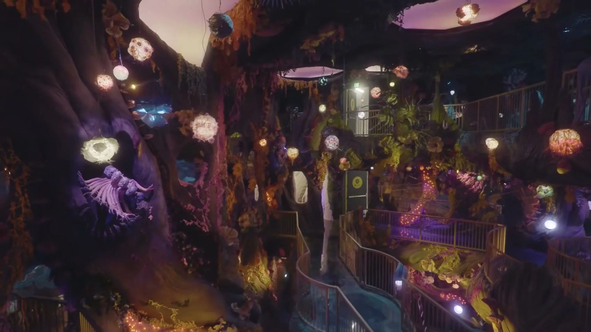 A wide shot of a fantastical, multi-level interior with glowing lights and organic structures, as a person walks down a winding ramp.