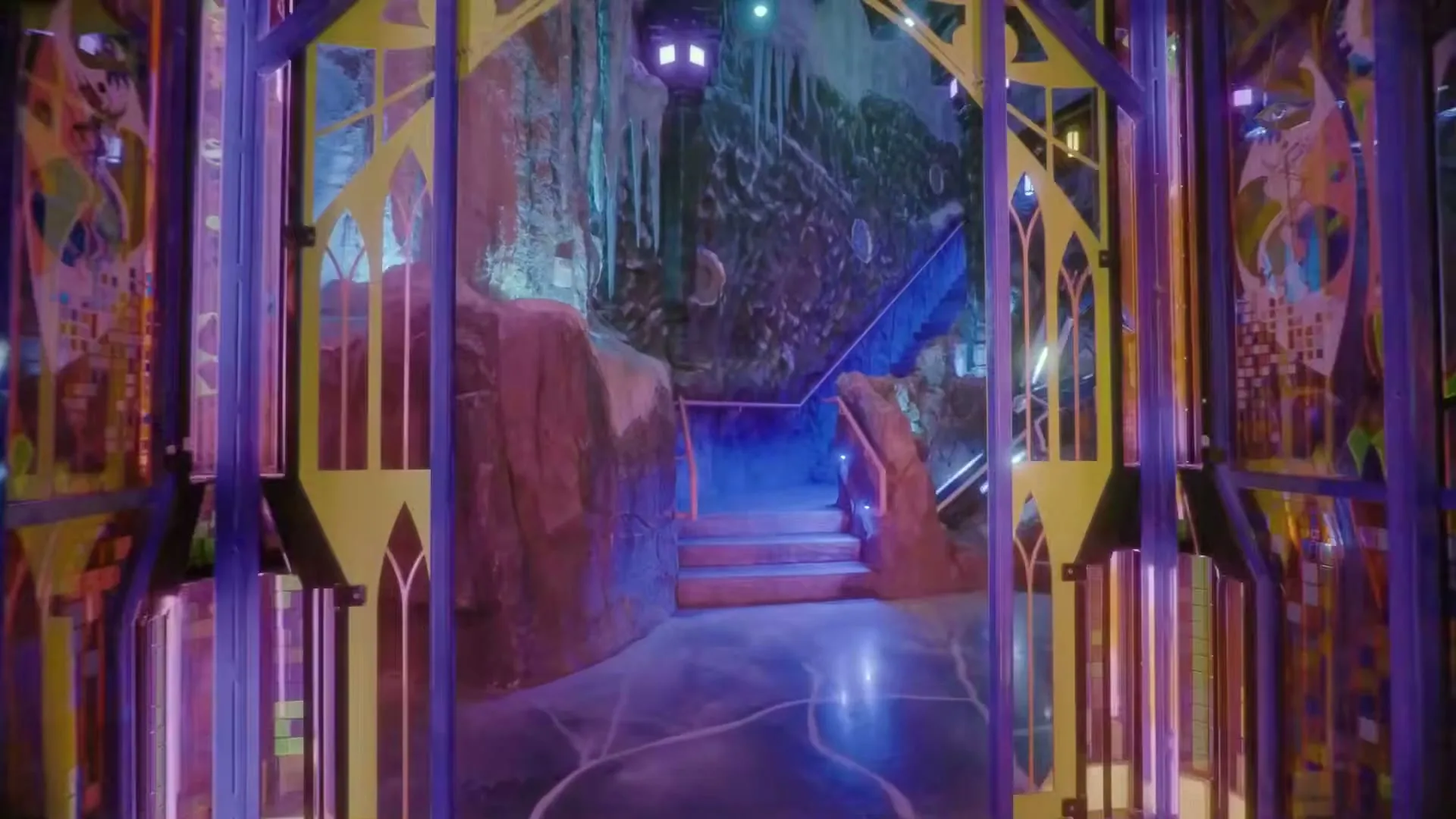 A wide shot of a futuristic, neon-lit hallway with glowing purple and yellow pillars, leading to a rocky cave-like area with stairs and a large, intricate robot.