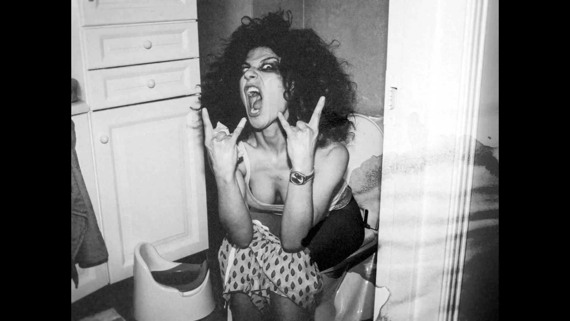 A black and white photo of a woman with big hair, wearing a patterned skirt, sitting on a toilet, screaming and making rock-and-roll hand gestures in a bathroom.