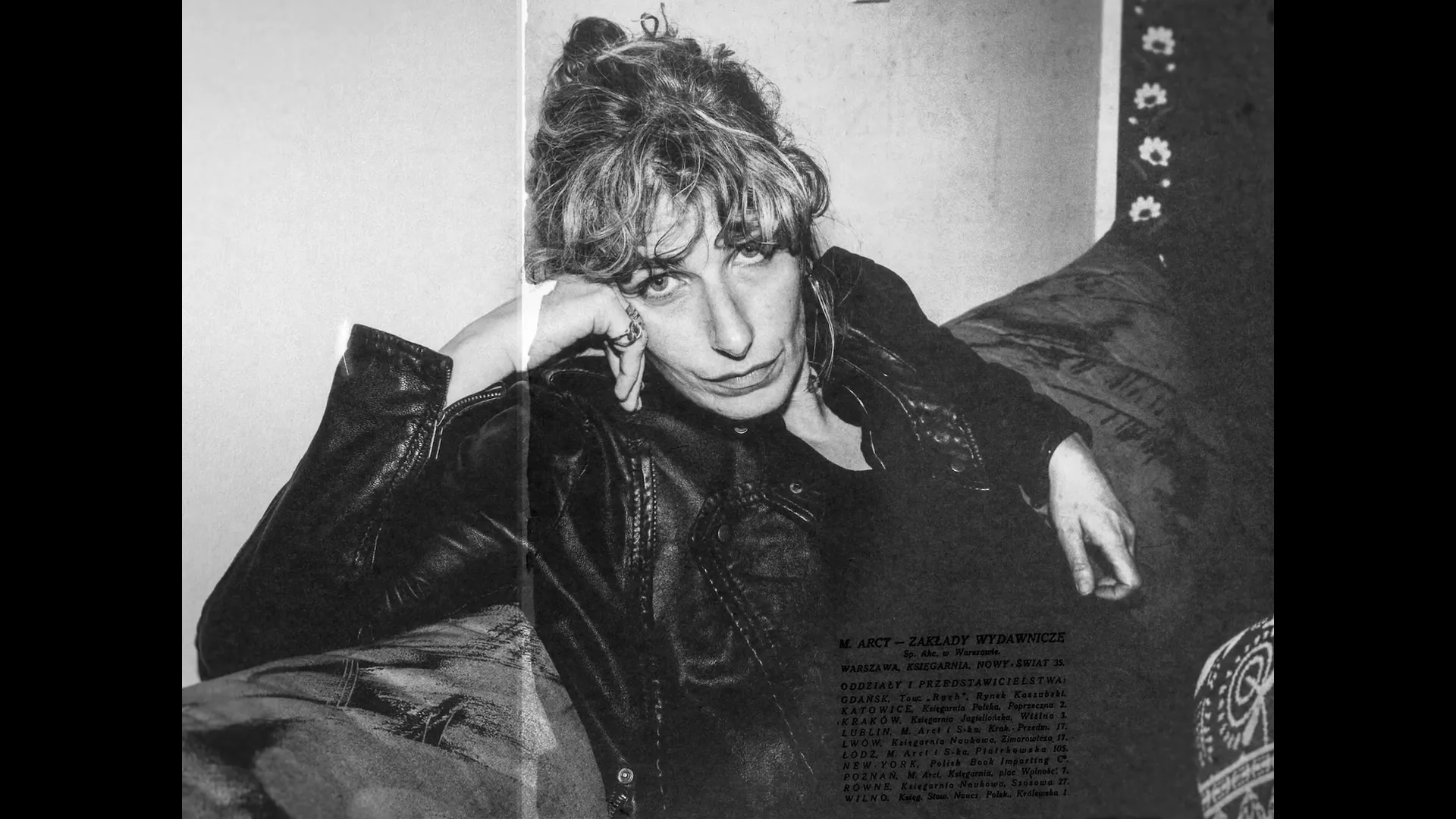 A woman with curly hair and a leather jacket lies on a couch, looking directly at the camera with a serious expression in a black and white shot.