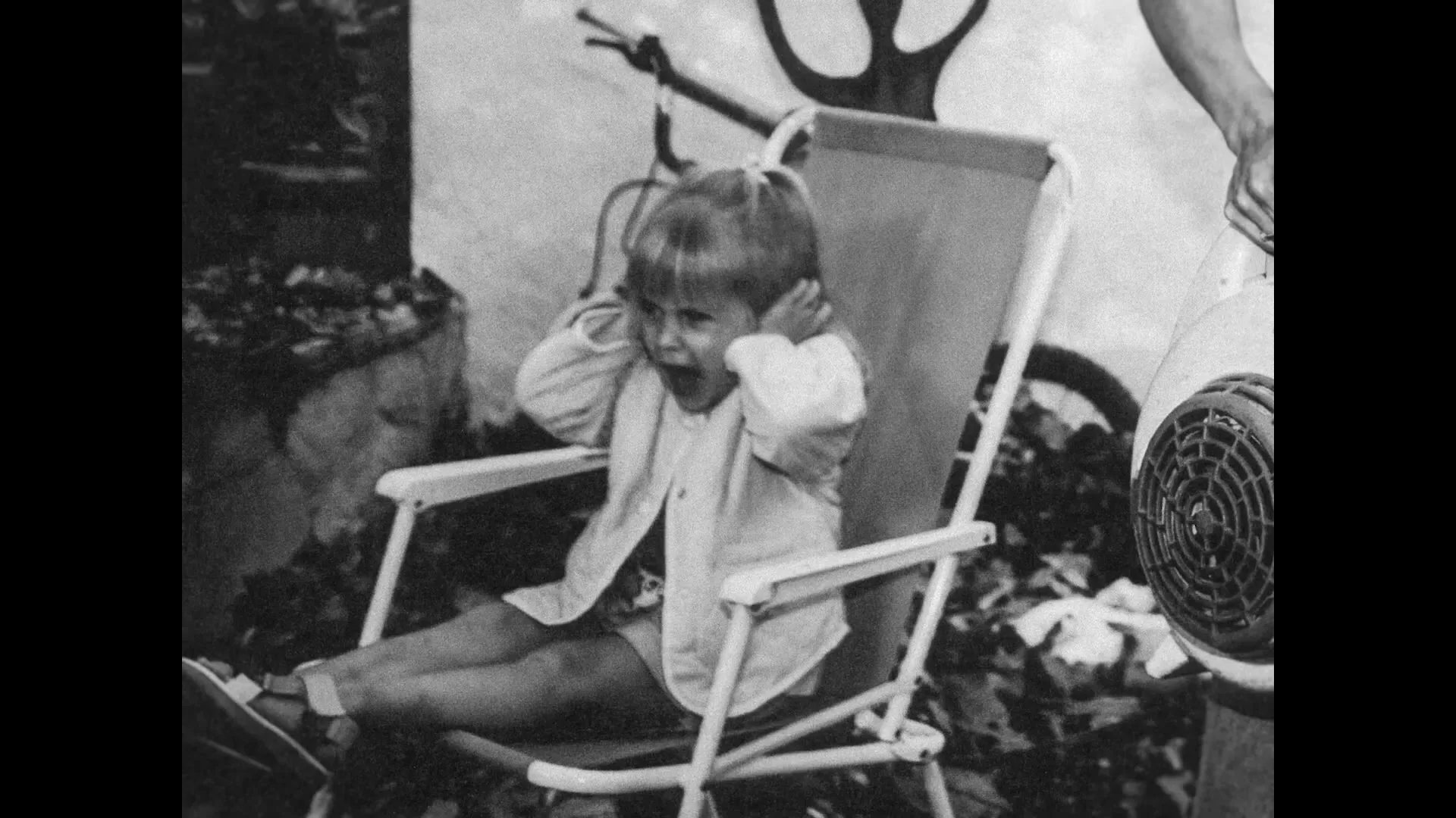 A young child with blonde hair sits in a folding chair, covering their ears and screaming as an adult holds a hairdryer nearby.