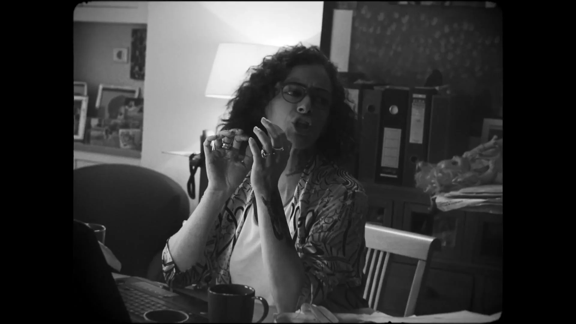 A woman with curly hair and glasses sits at a table, talking and gesturing with her hands, in a home setting.