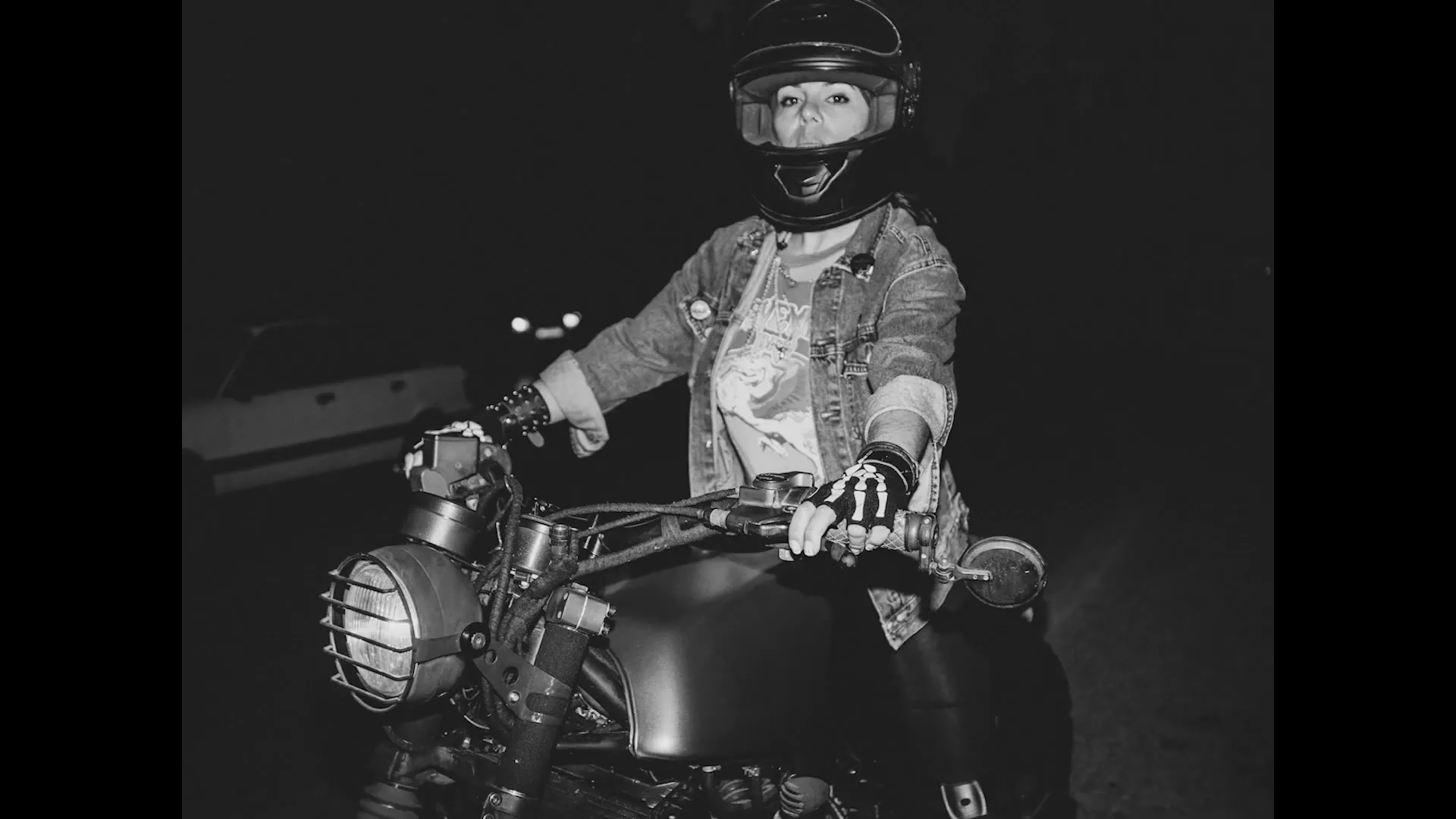 A woman in her 20s, wearing a helmet and denim jacket, sits on a motorcycle at night, looking directly at the camera.
