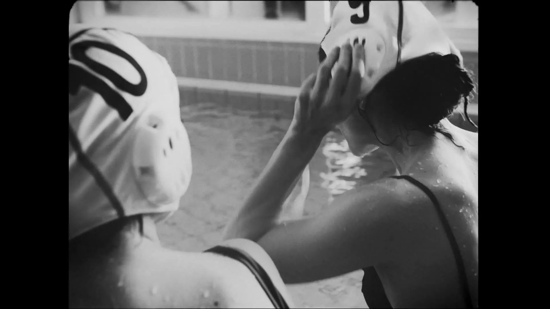 Two women in water polo caps and swimsuits are in a swimming pool, one adjusting her cap, in a black and white shot.