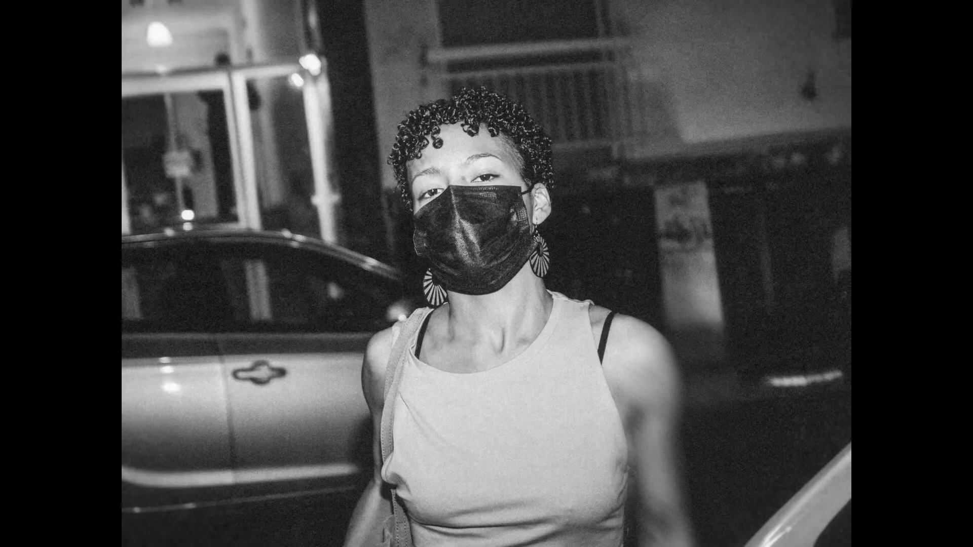 A young woman with curly hair and a face mask looks directly at the camera on a city street at night.