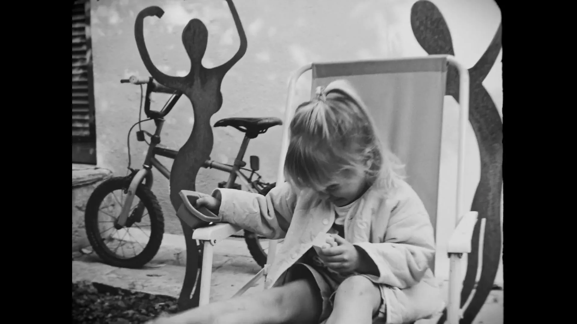 A young girl with blonde hair sits in a folding chair, looking down at her hands, with a bicycle and abstract sculptures in the background.