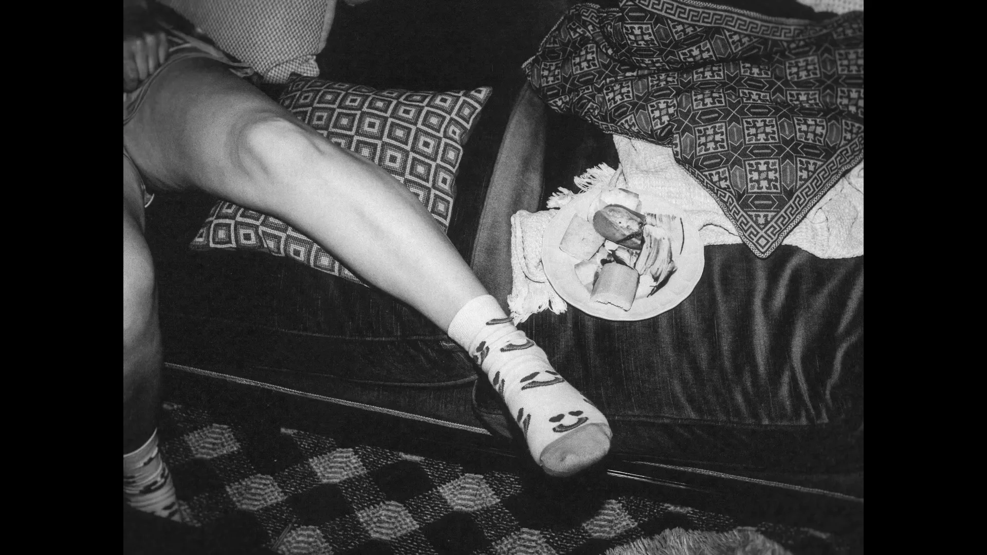 A person's leg with a patterned sock rests on a couch next to a plate of food, captured in a black and white, grainy shot.