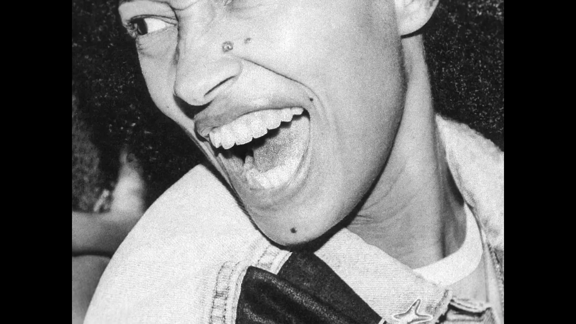 A young woman with curly hair, wearing a denim jacket, laughs joyfully in a black and white extreme closeup.