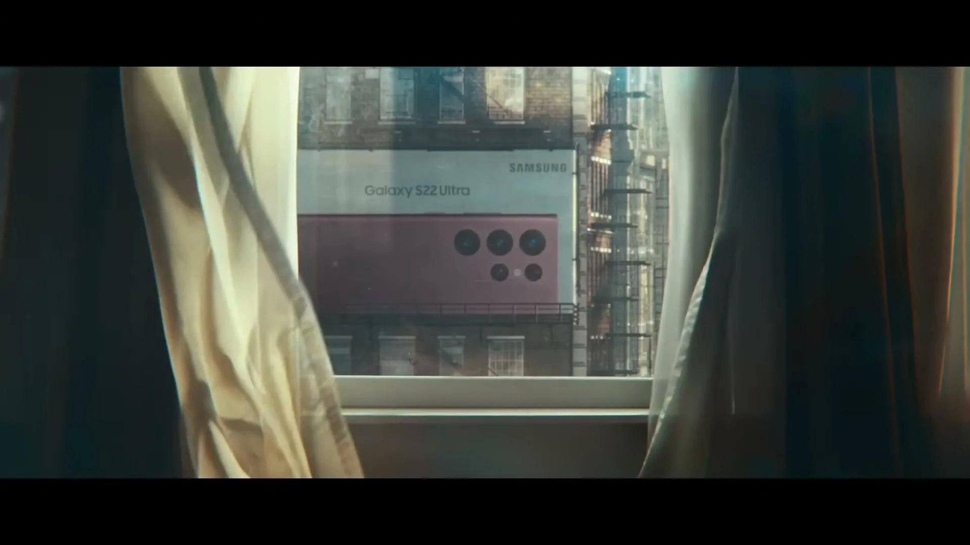 A window with white curtains reveals a large billboard outside, displaying a pink Samsung Galaxy S22 Ultra phone in an urban setting.