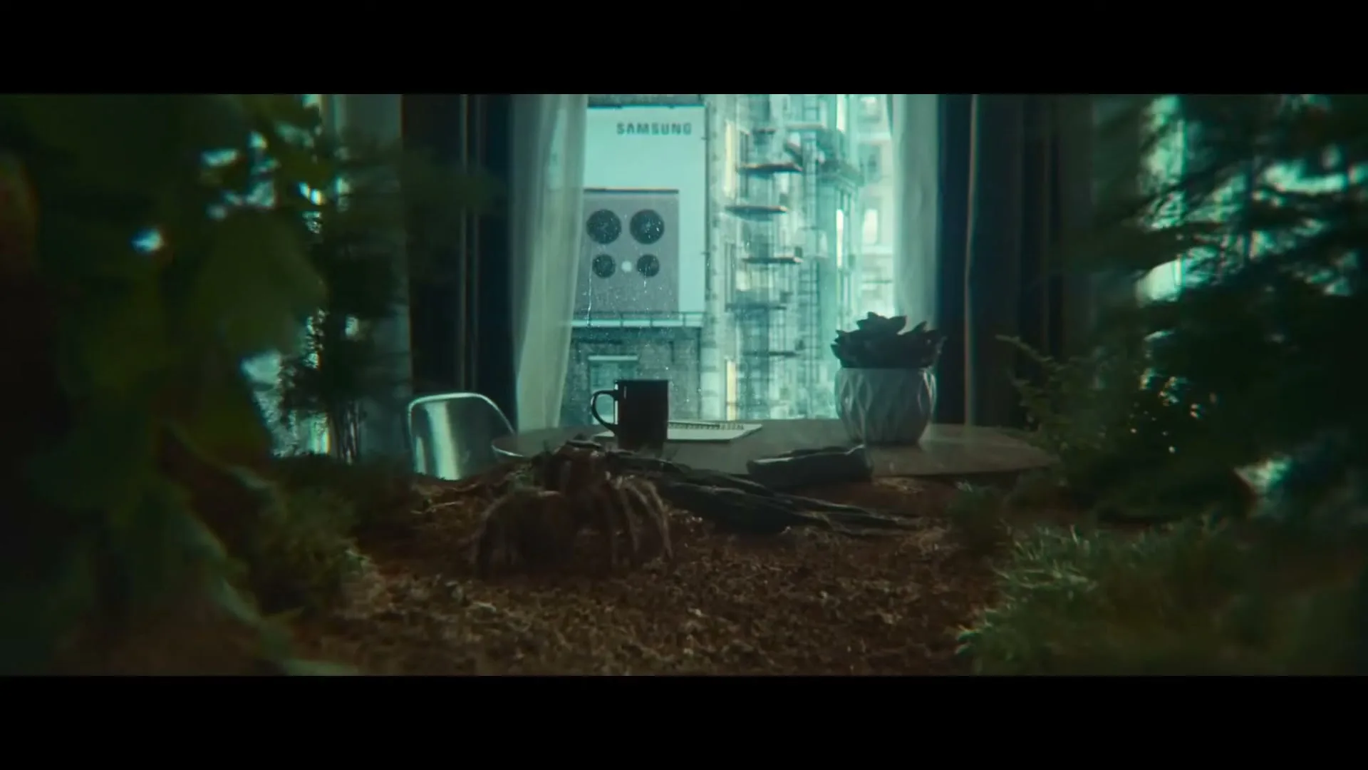 A tarantula crawls on a desk covered in dirt and plants, with a city view and a Samsung billboard visible through a window.