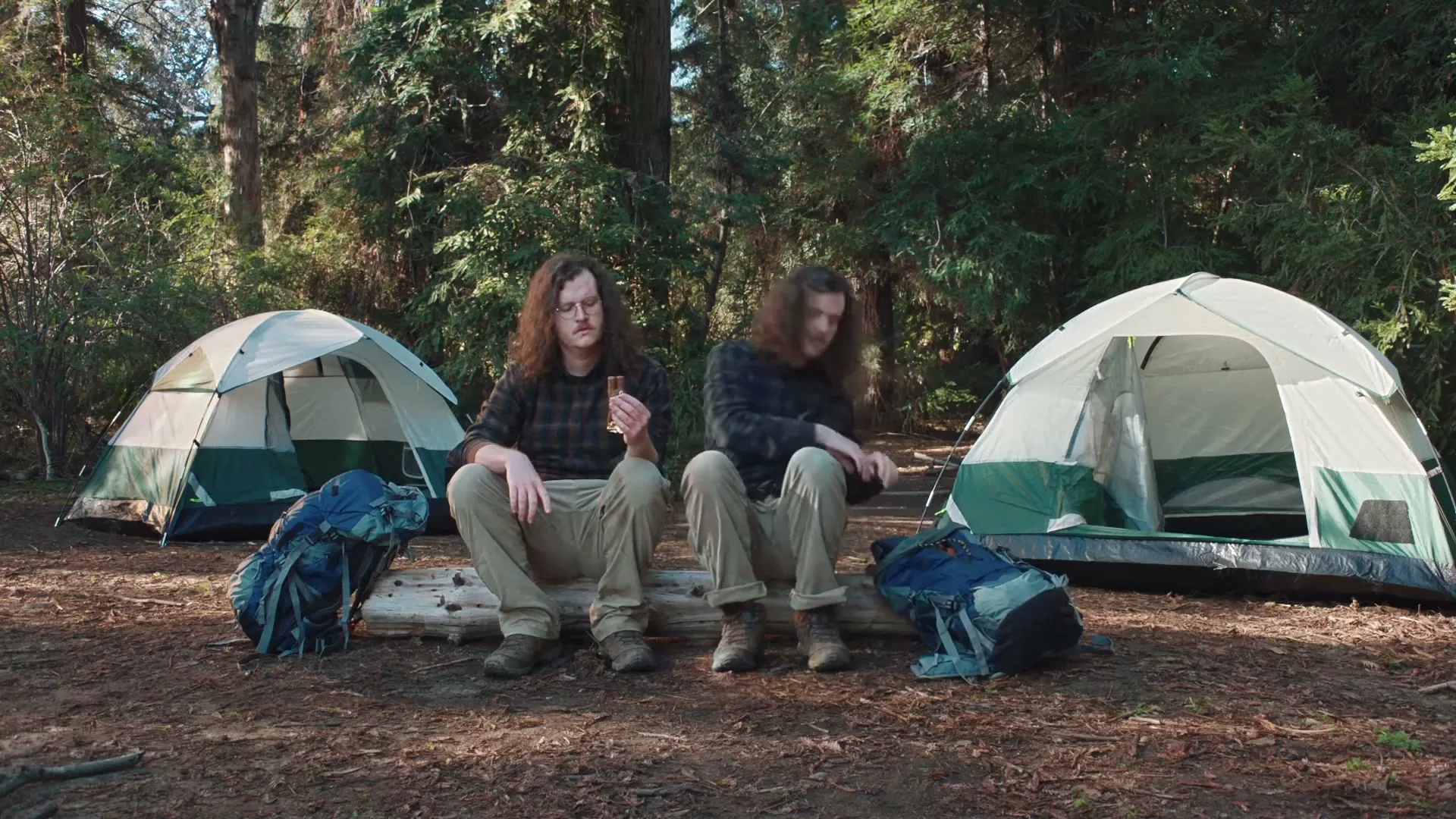 Two men with long curly hair and glasses sit on a log between two tents in a forest, one holding a Twix bar.