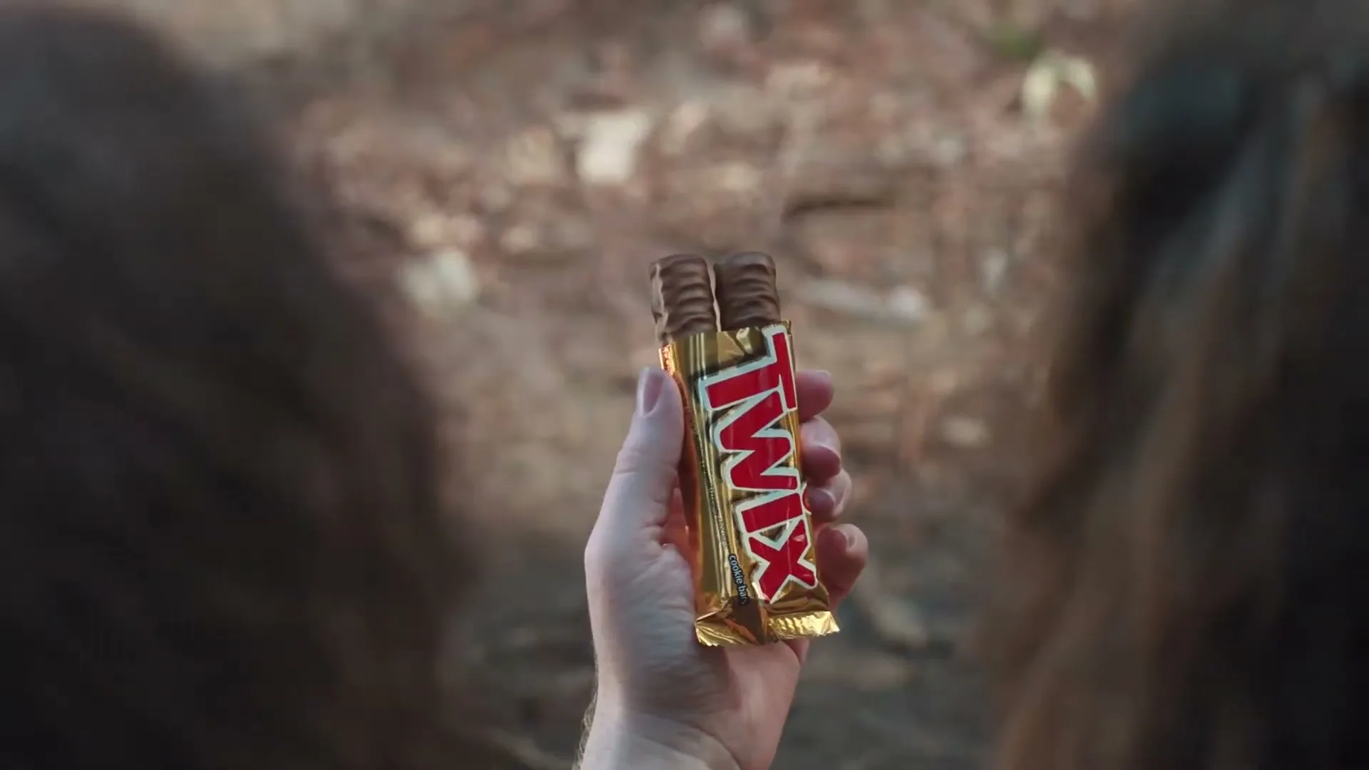 A person's hand holds up a Twix bar, framed between two blurry, dark, furry objects, possibly bear ears, in an outdoor setting.