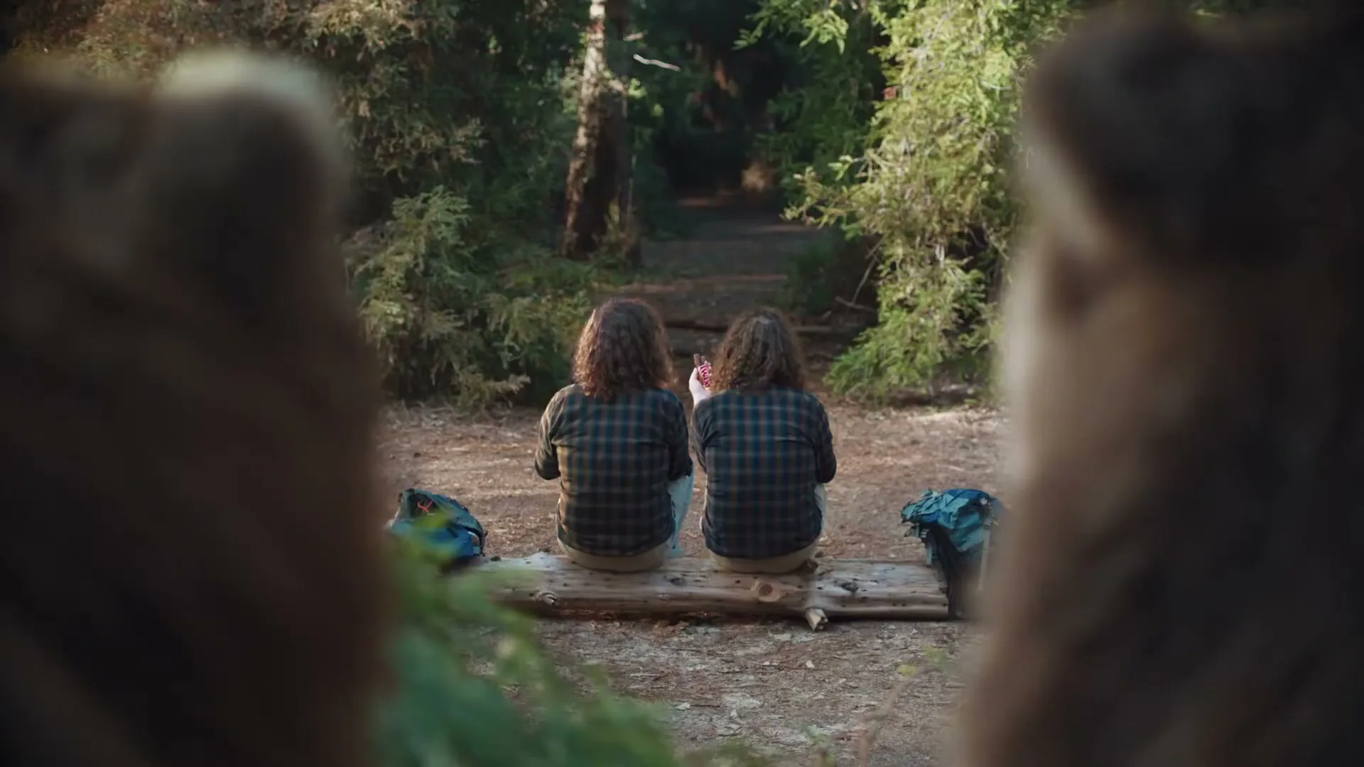 Two people with long, curly hair sit on a log in a forest, seen from behind through a blurry foreground.