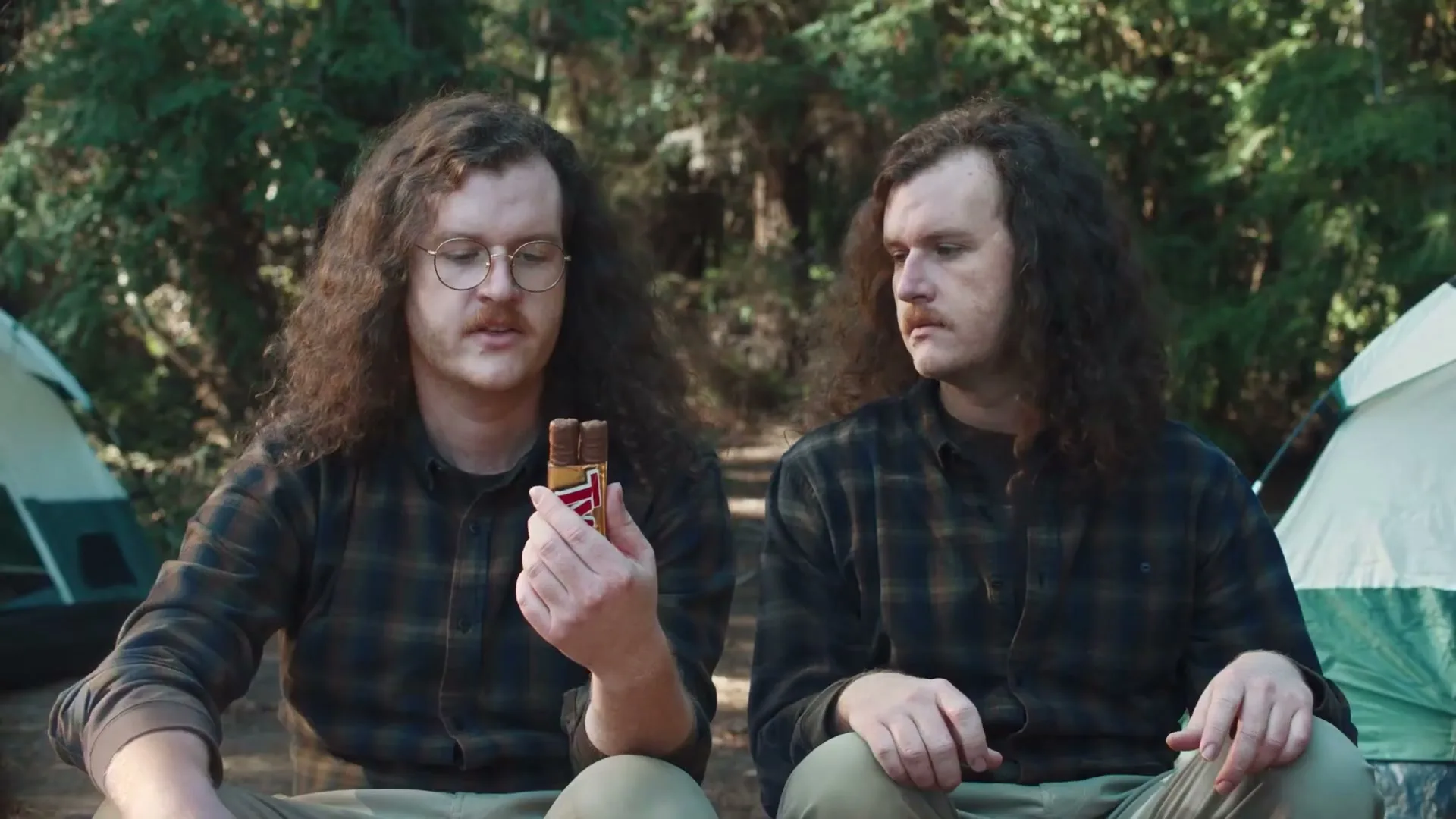 Two men with long curly hair and mustaches sit in a forest campsite, one holding a Twix bar while the other looks on.
