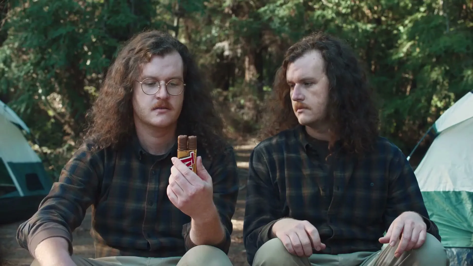 Two men with long curly hair and mustaches sit in a forest campsite, one holding a Twix bar, looking at it intently while the other looks on.