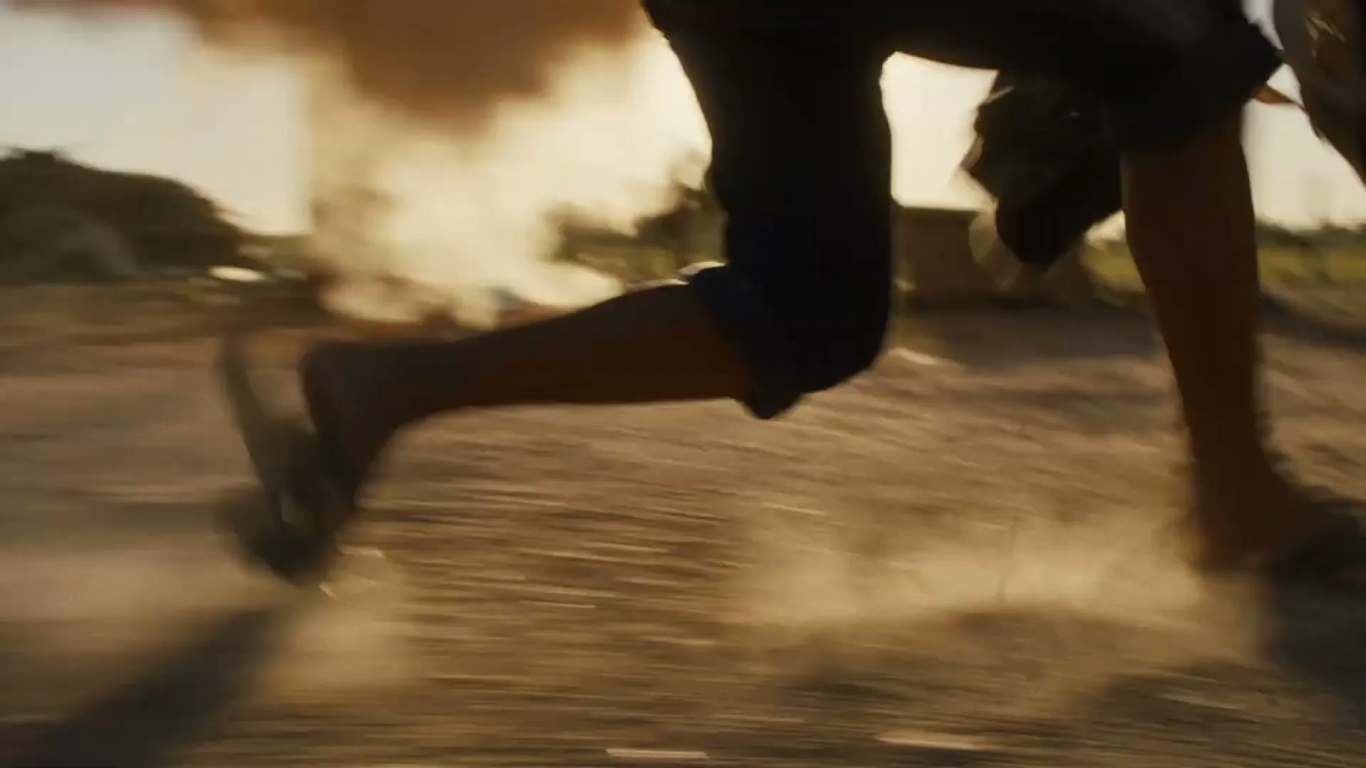 A person in dark shorts and sandals runs across a dusty field, kicking up dirt as an explosion happens behind them under a bright sky.