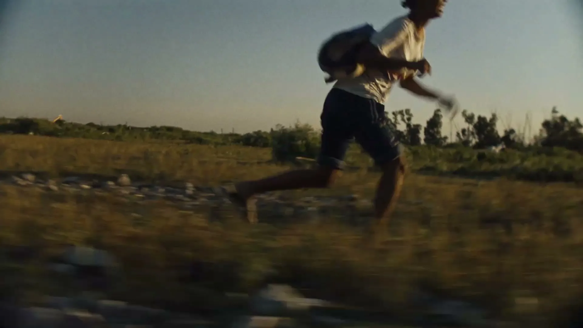 A young male refugee, wearing a white t-shirt and shorts with a backpack, runs barefoot across a dry, grassy field under a clear sky.
