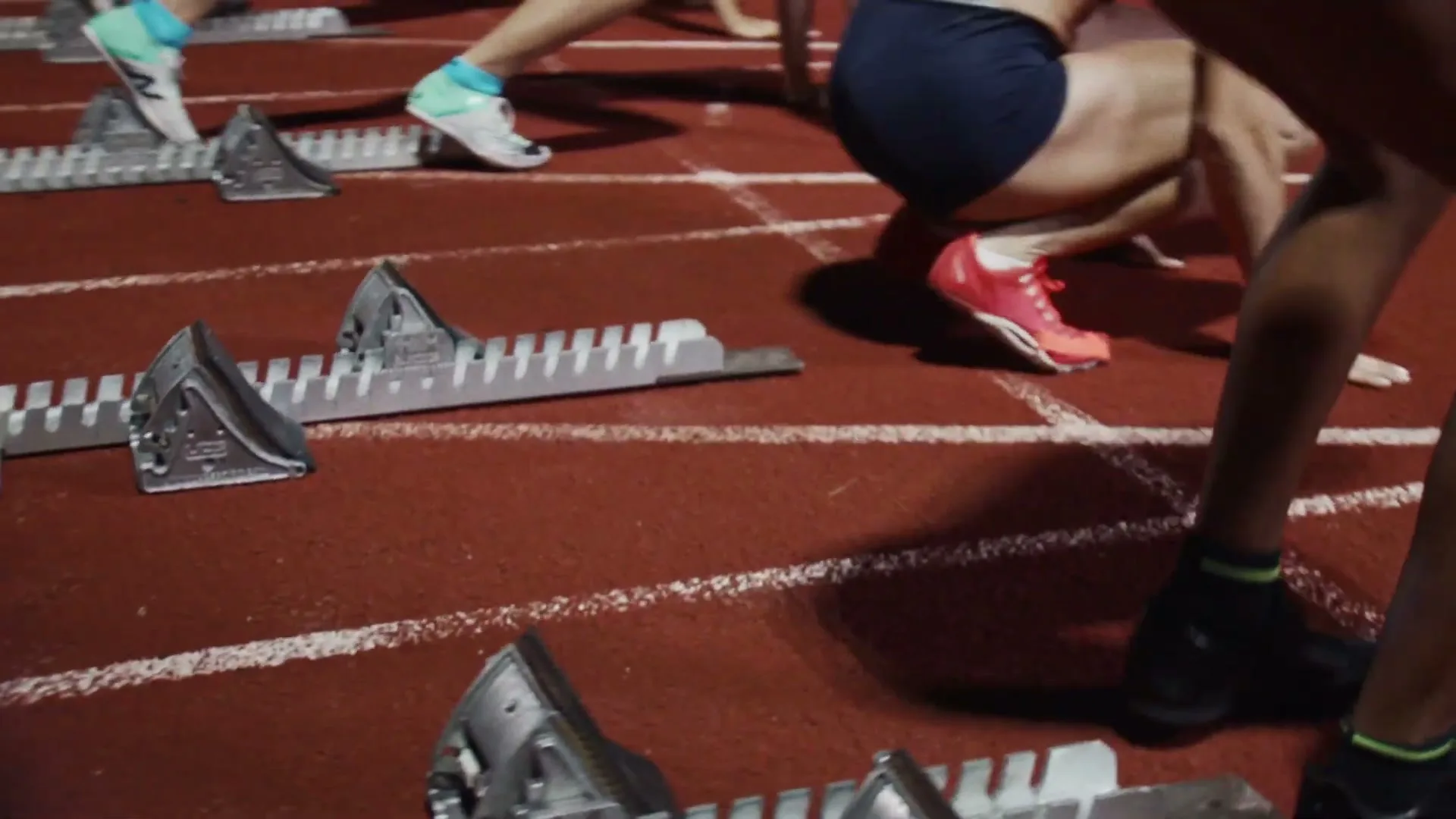 A group of athletes in running shoes and athletic wear get into position on starting blocks on a red track at night.