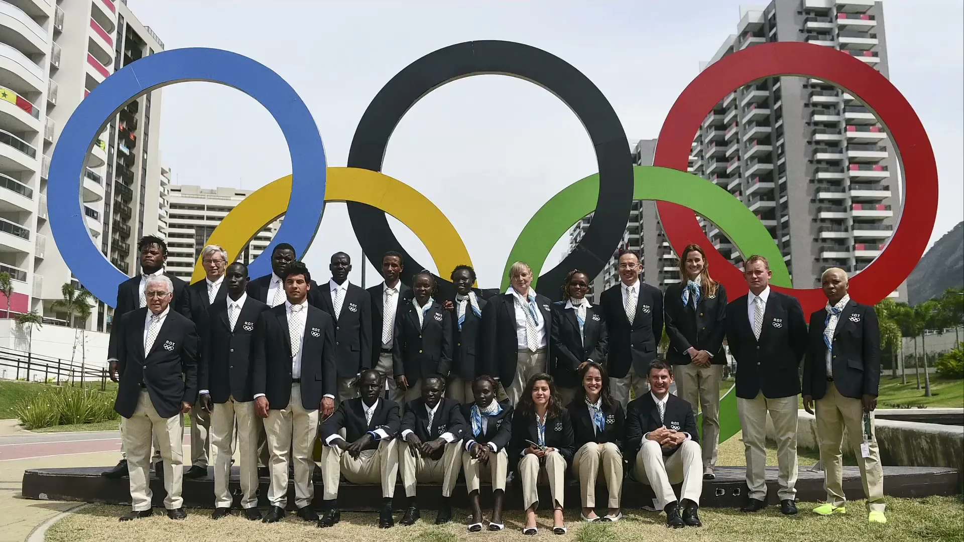 A large group of diverse people, dressed in matching blazers and light-colored pants, pose for a group photo in front of a large Olympic rings sculpture outdoors.