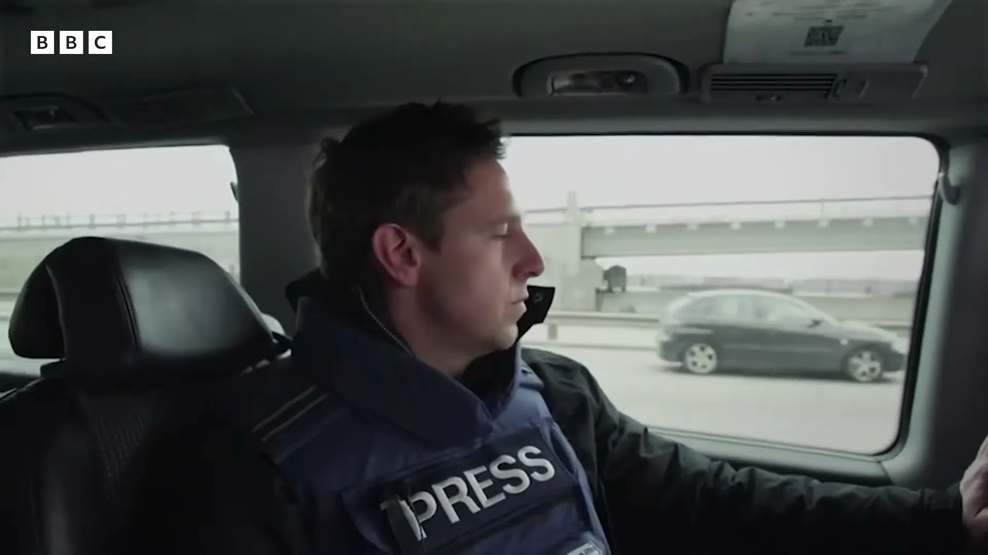 A male journalist in a "PRESS" vest looks out the window of a moving car, observing a highway overpass and passing traffic.