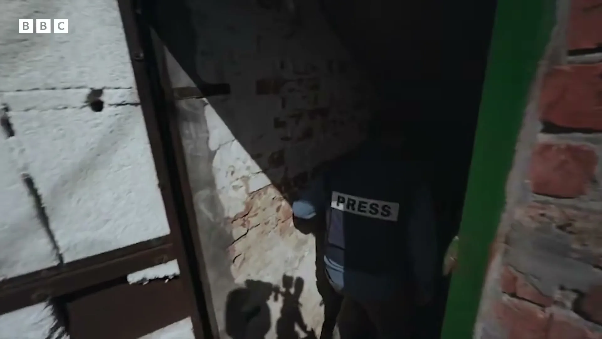 A person wearing a "PRESS" vest walks through a narrow, dark doorway in a dilapidated building, with brick and concrete walls visible.