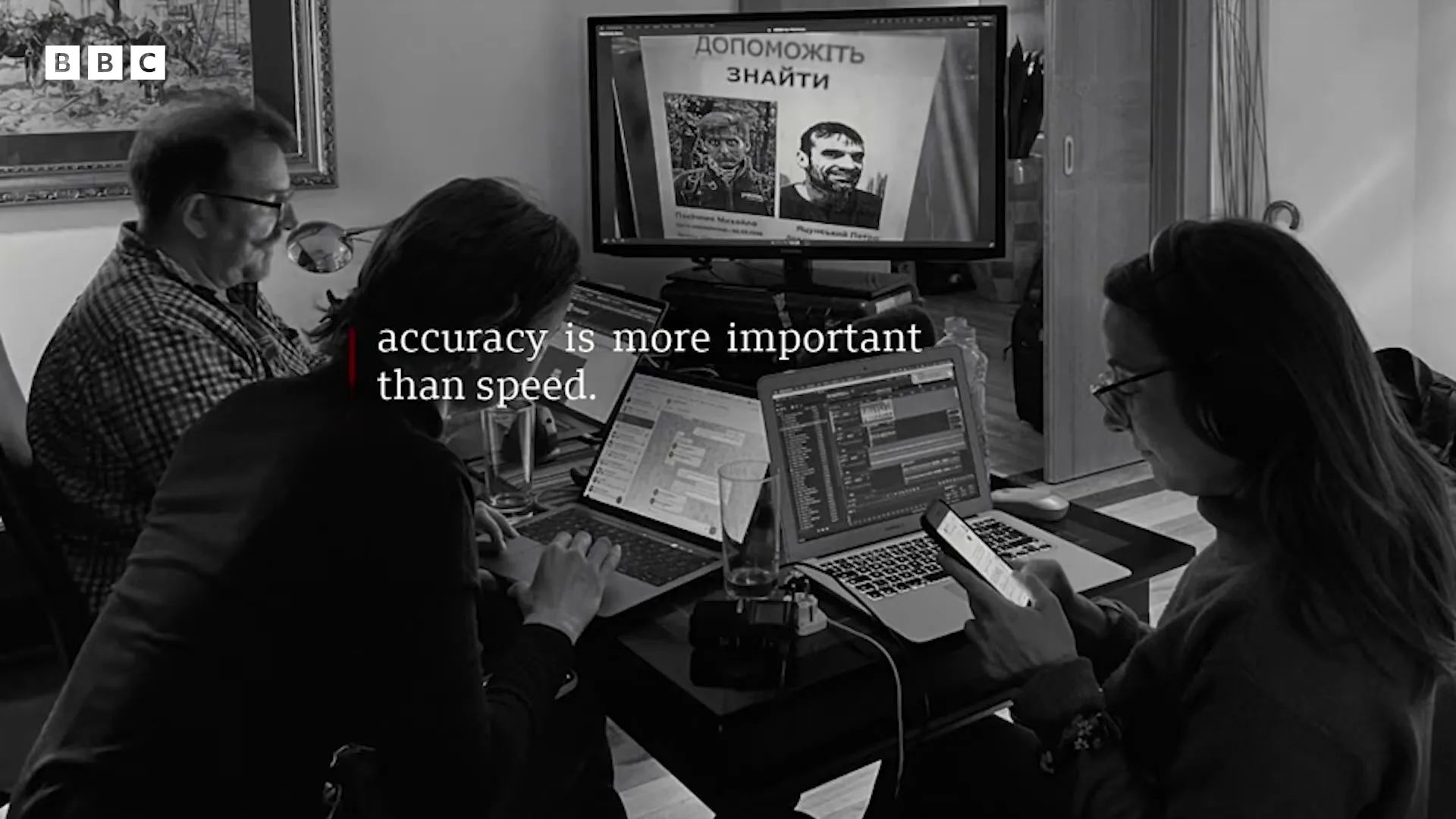 Three people, two women and one man, are working on laptops and a phone at a table, with a monitor displaying a missing person poster in the background.