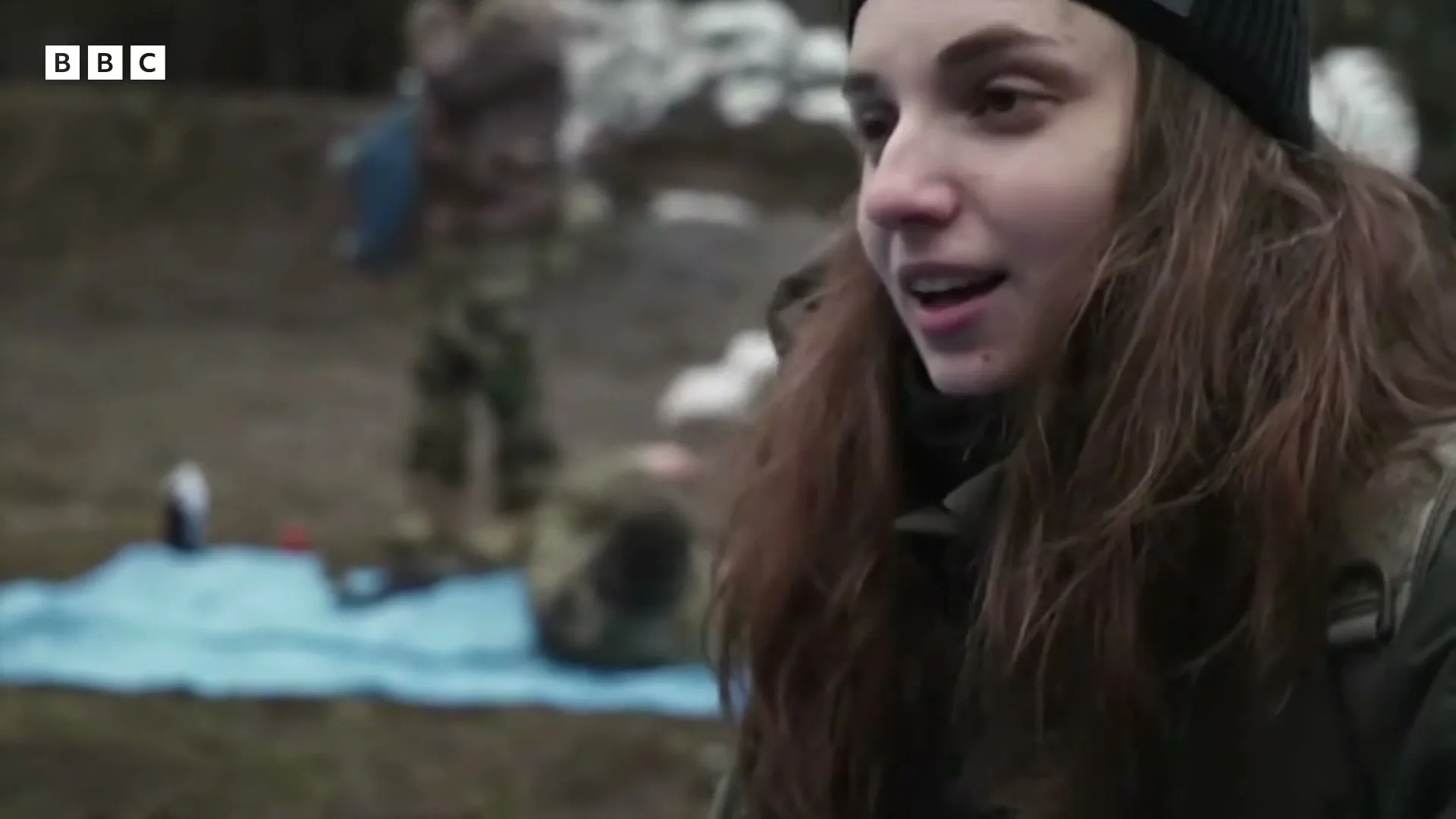 A young woman with long brown hair and a black beanie smiles and talks, while people in camouflage are blurred in the background.