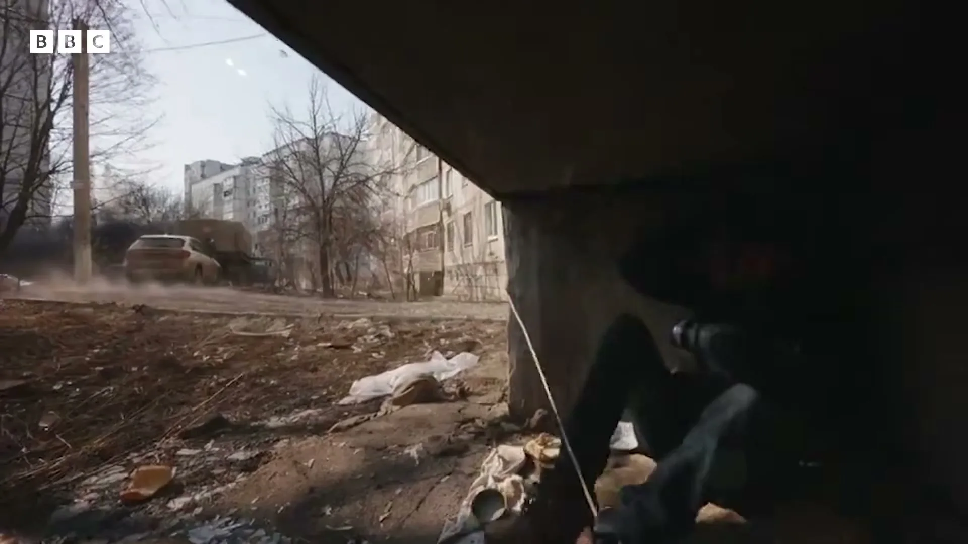A person in dark clothing crouches under a concrete slab, filming a car driving on a dusty road in a war-torn urban area.
