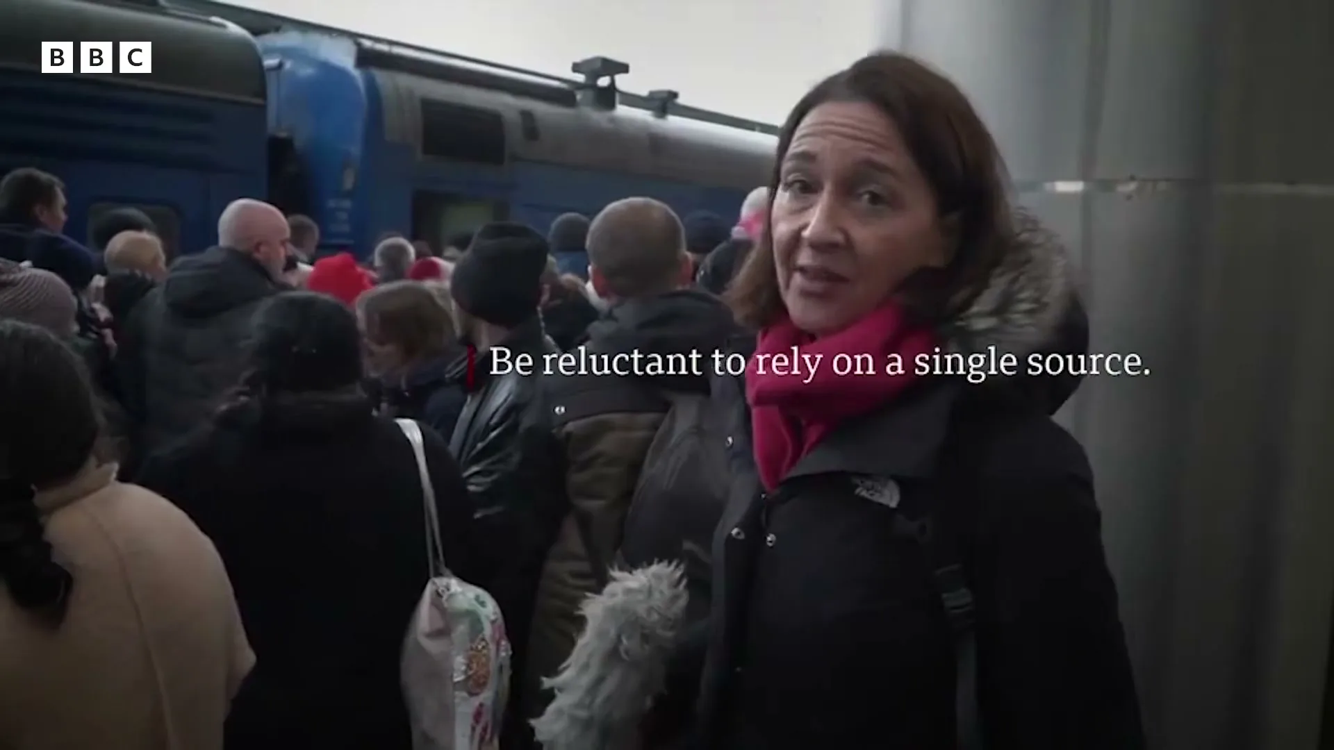 A female reporter in a black winter coat and pink scarf speaks to the camera amidst a crowd of people and a blue train in the background.
