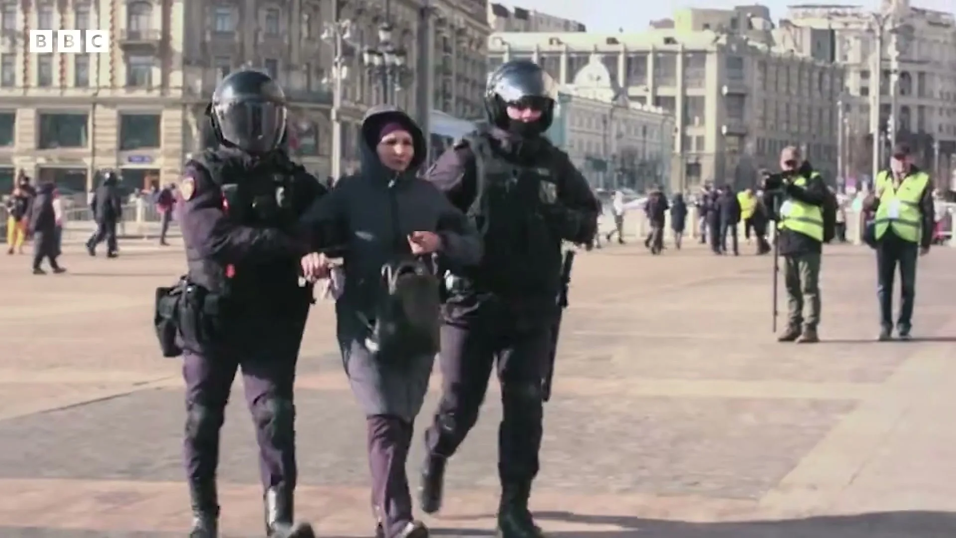 Two police officers in riot gear escort a woman in a dark coat and headscarf across a sunny city square, with other people in the background.