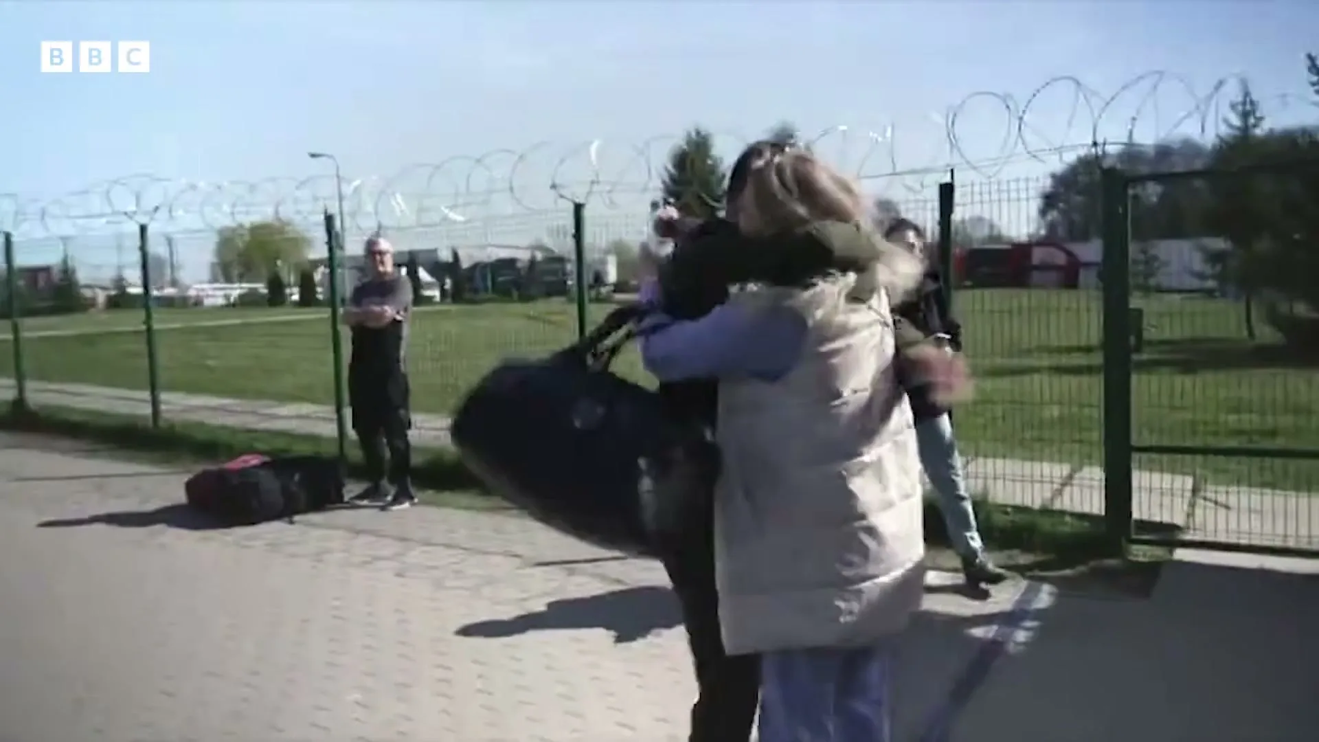 A man and a woman embrace warmly near a barbed wire fence, while another man stands by with luggage in an outdoor setting.