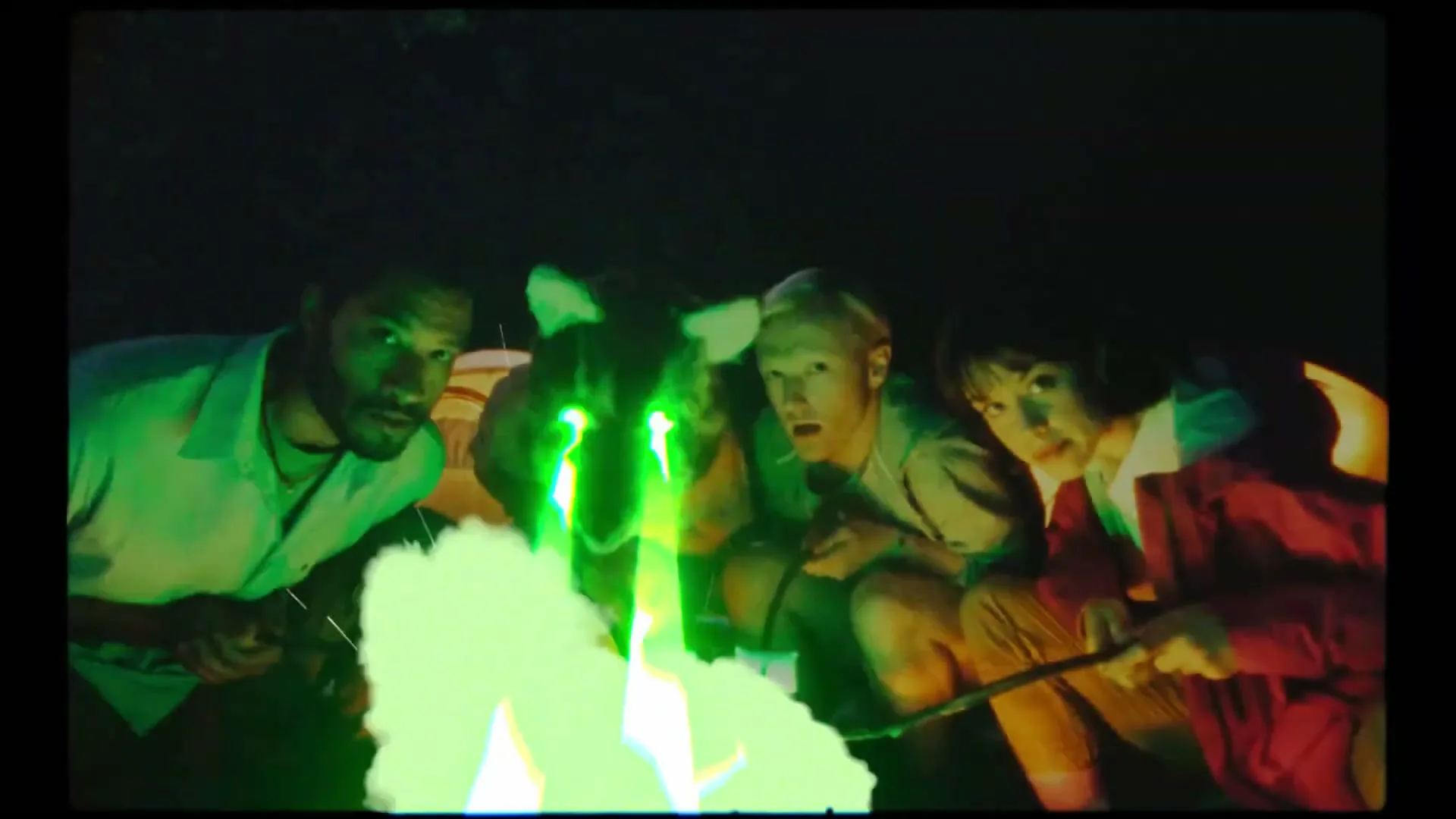 Three young adults and a mascot sit around a campfire, roasting marshmallows, with green light emanating from the fire and mascot's eyes.
