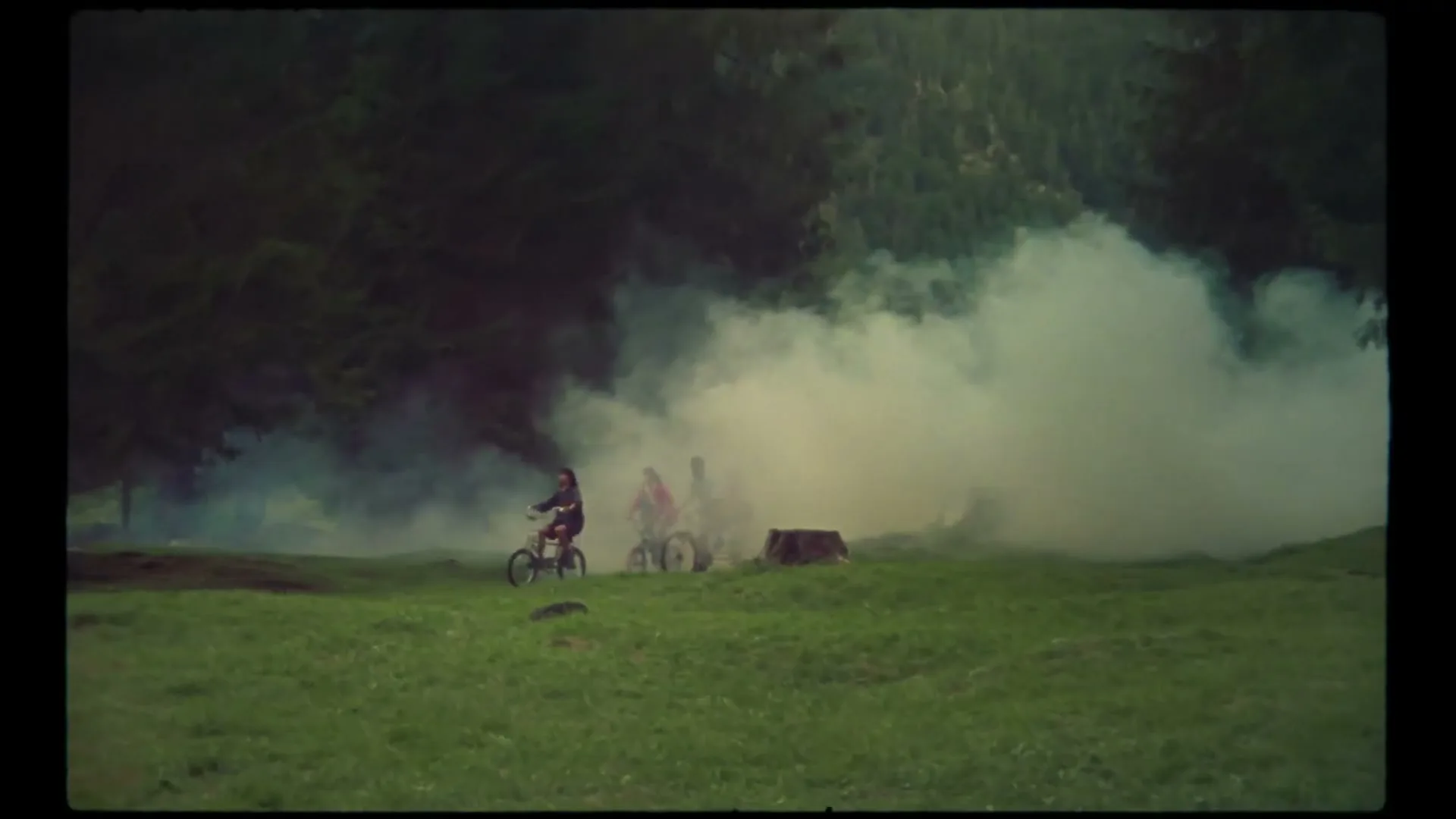 Four young people ride BMX bikes through a grassy field with smoke in the background, surrounded by trees.