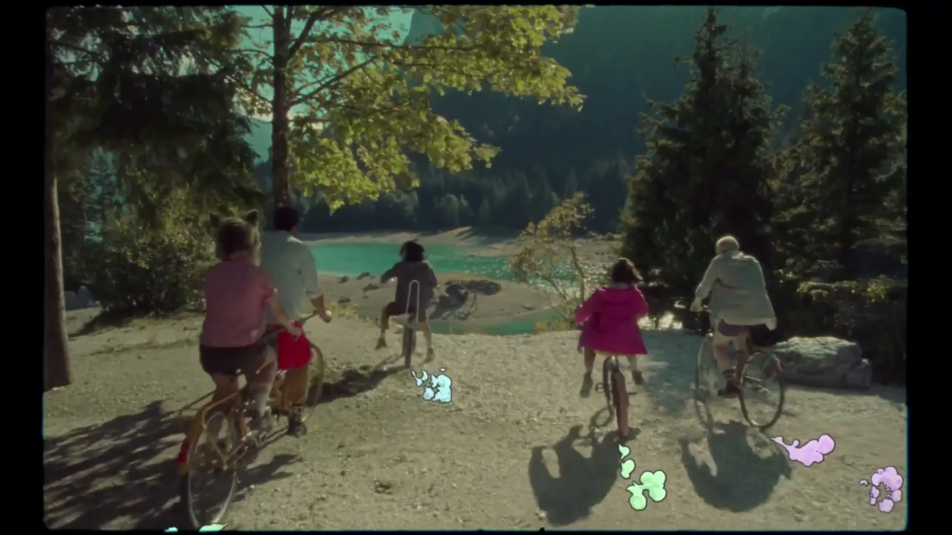 Four people of various ages ride bicycles down a dirt path towards a bright blue lake surrounded by mountains and trees on a sunny day.