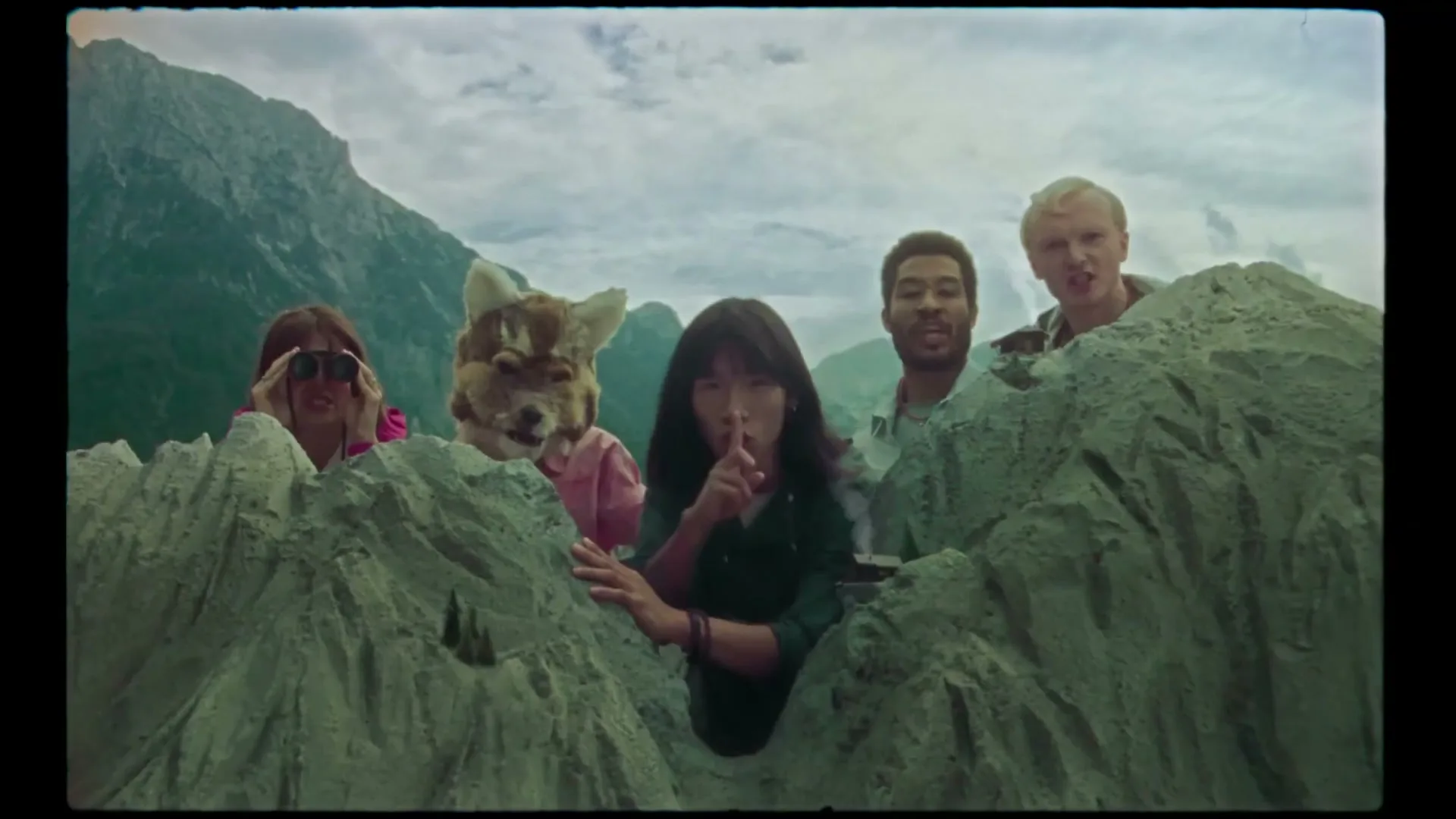 Five people, one in a fox mask, hide behind miniature mountains, looking surprised and one shushing, with a large mountain range in the background under a cloudy sky.